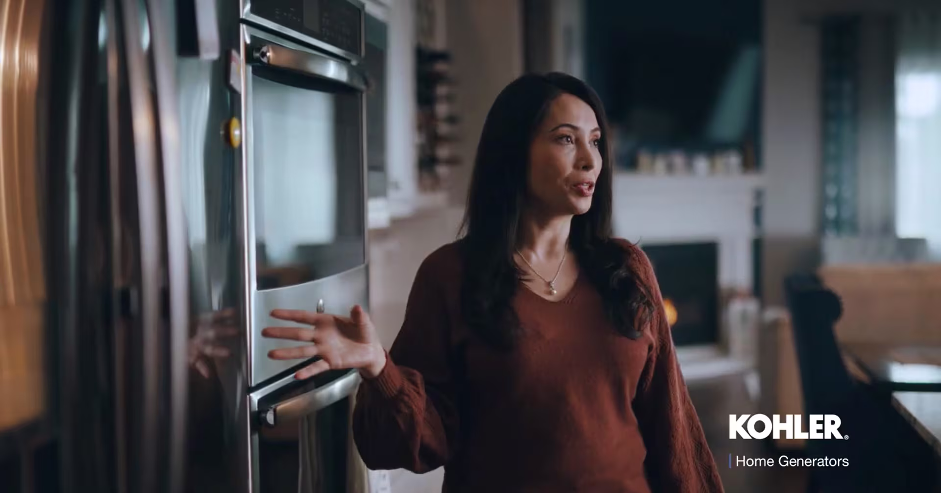 Long haired woman, wearing a brown blouse, in front of the appliances in her kitchen, talking to someone off-camera.