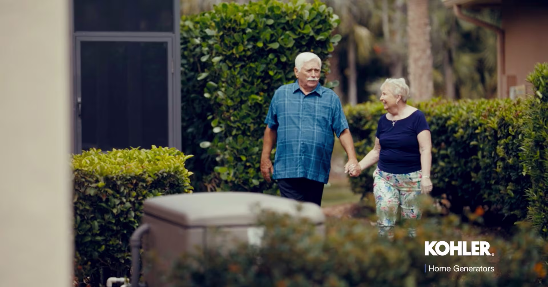 Elderly couple, walking holding hands through their yard in front of a Kohler home generator.