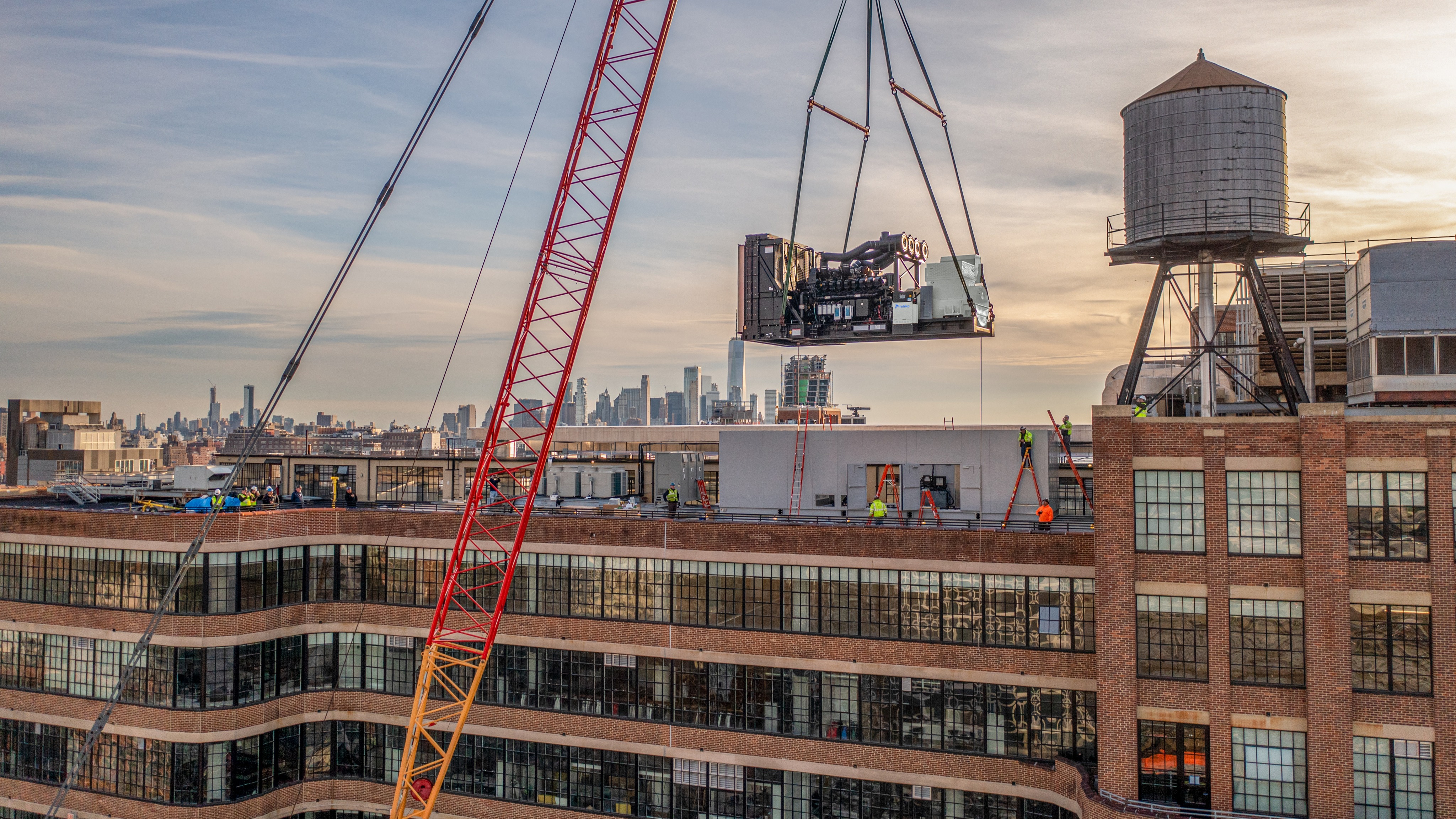 Crane lifting a diesel generator to the rooftop of a building under a warm late afternoon sky.