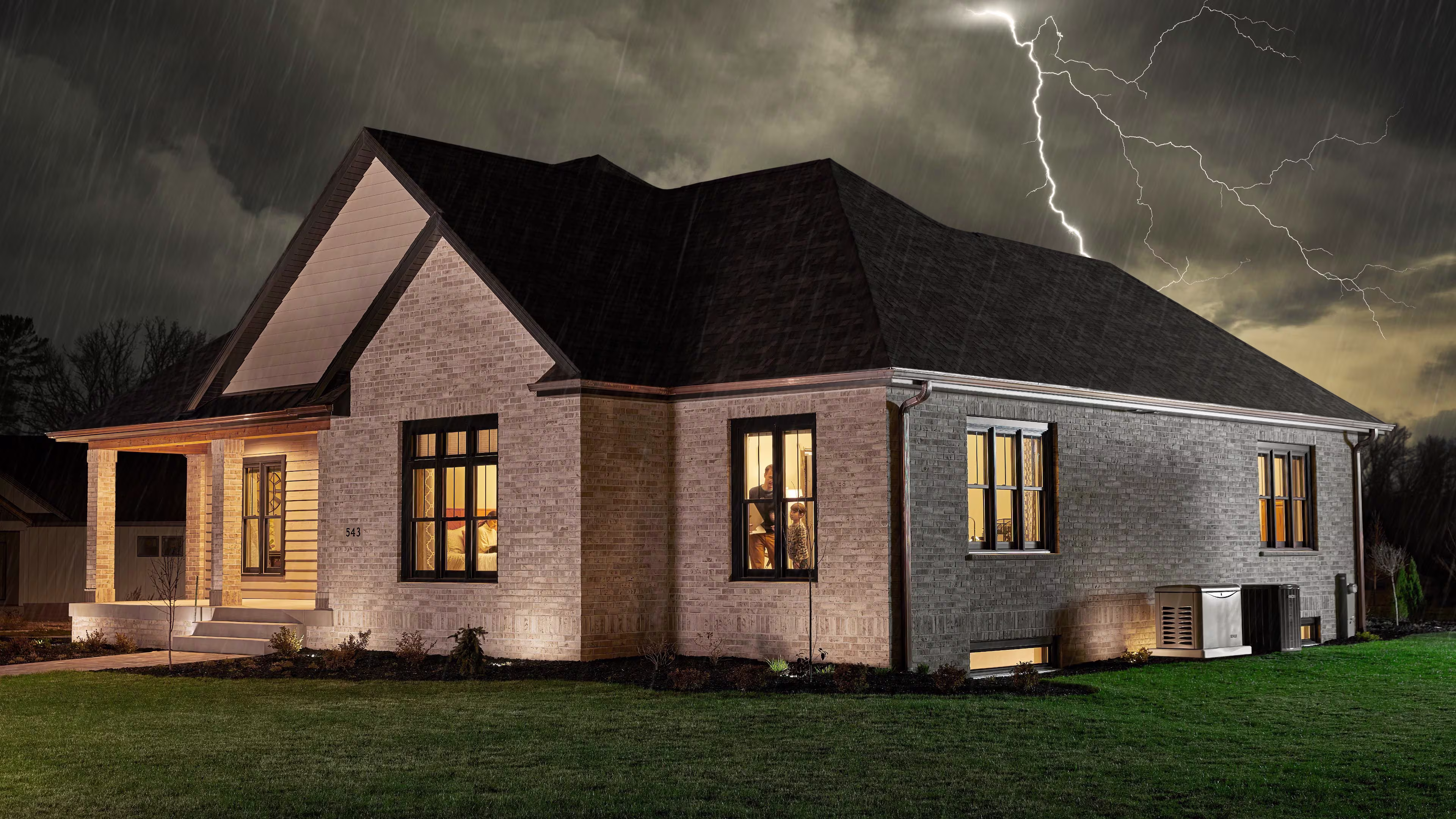 A large brick house with a Kohler generator along a wall in a residential neighborhood at night with a storm and lightning outside and the lights on inside 