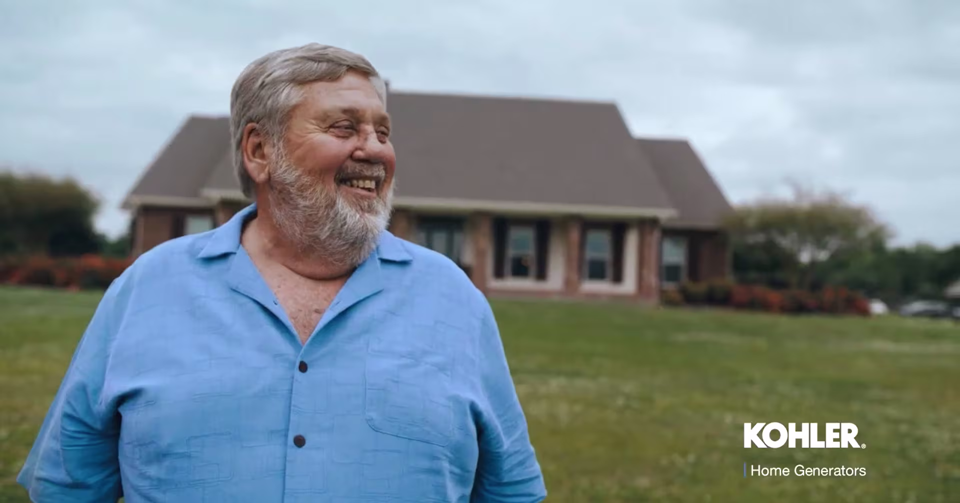 White haired man wearing a blue shirt and smiling on a yard, in the background there's a brown house.