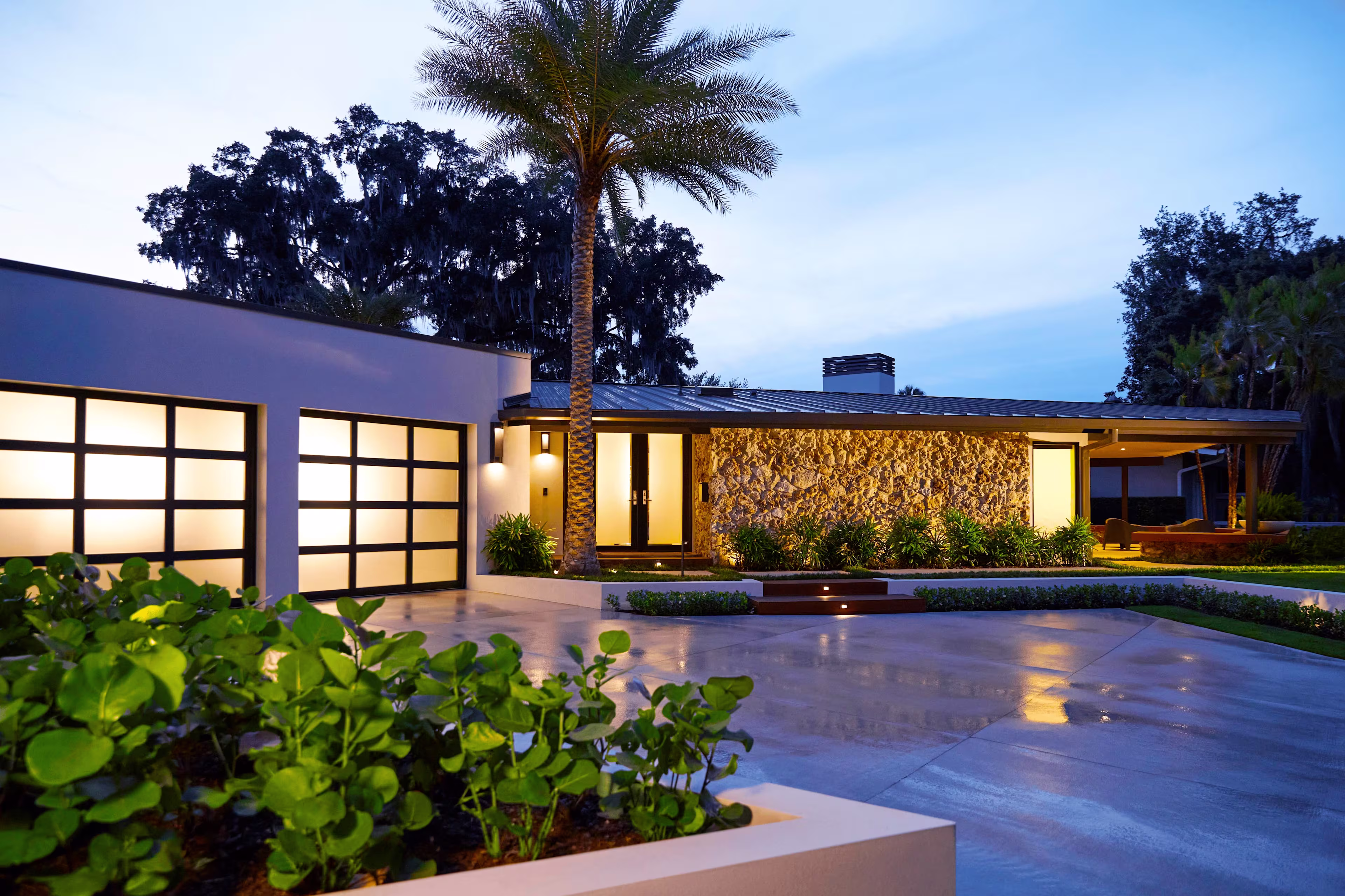 Front view of a house with its indoor lights on featuring a palm tree and a garden in the yard.