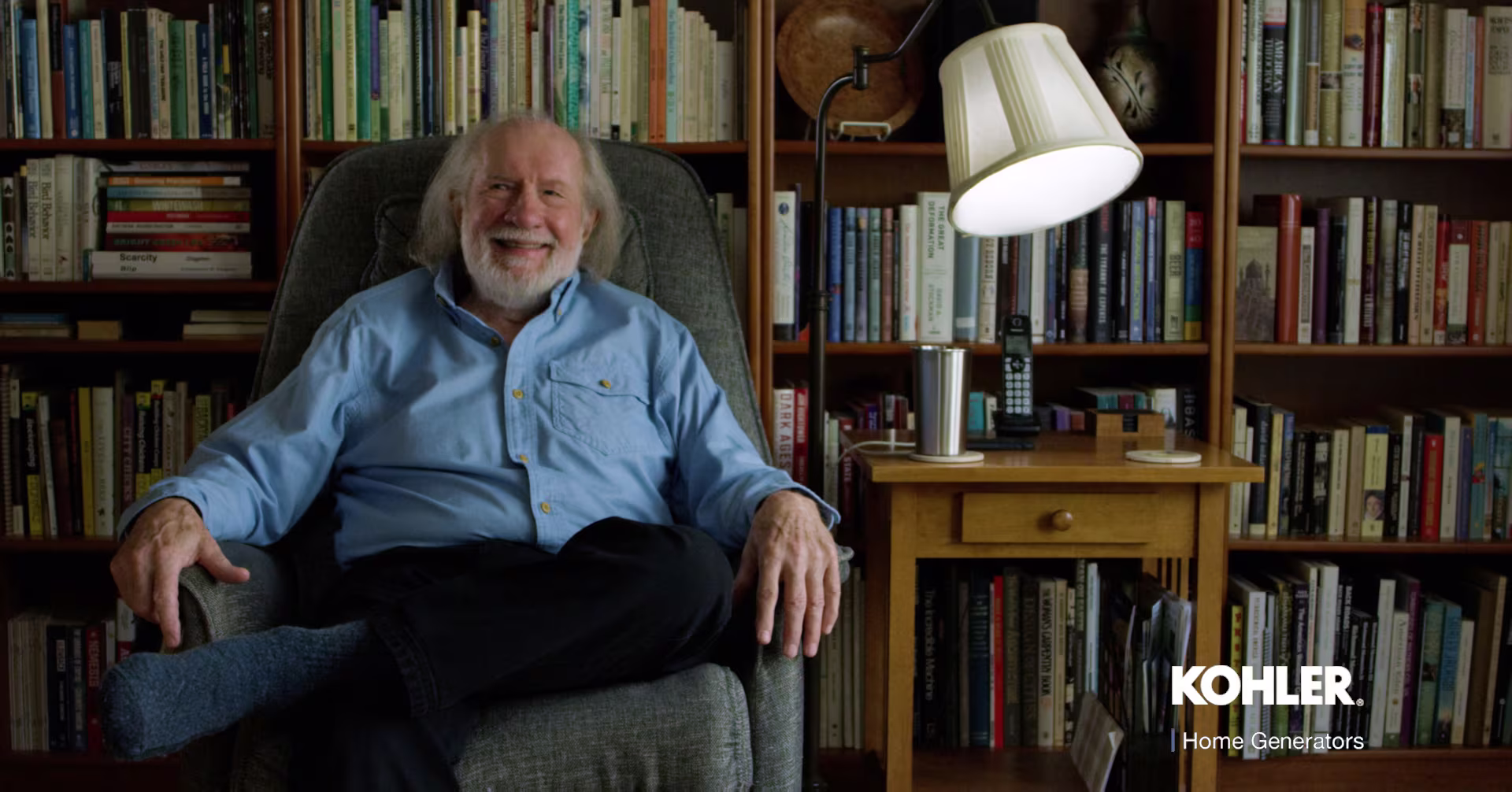 Elderly man wearing a blue shirt and dark pants, comfortably sitting on a grey chair in a home library.