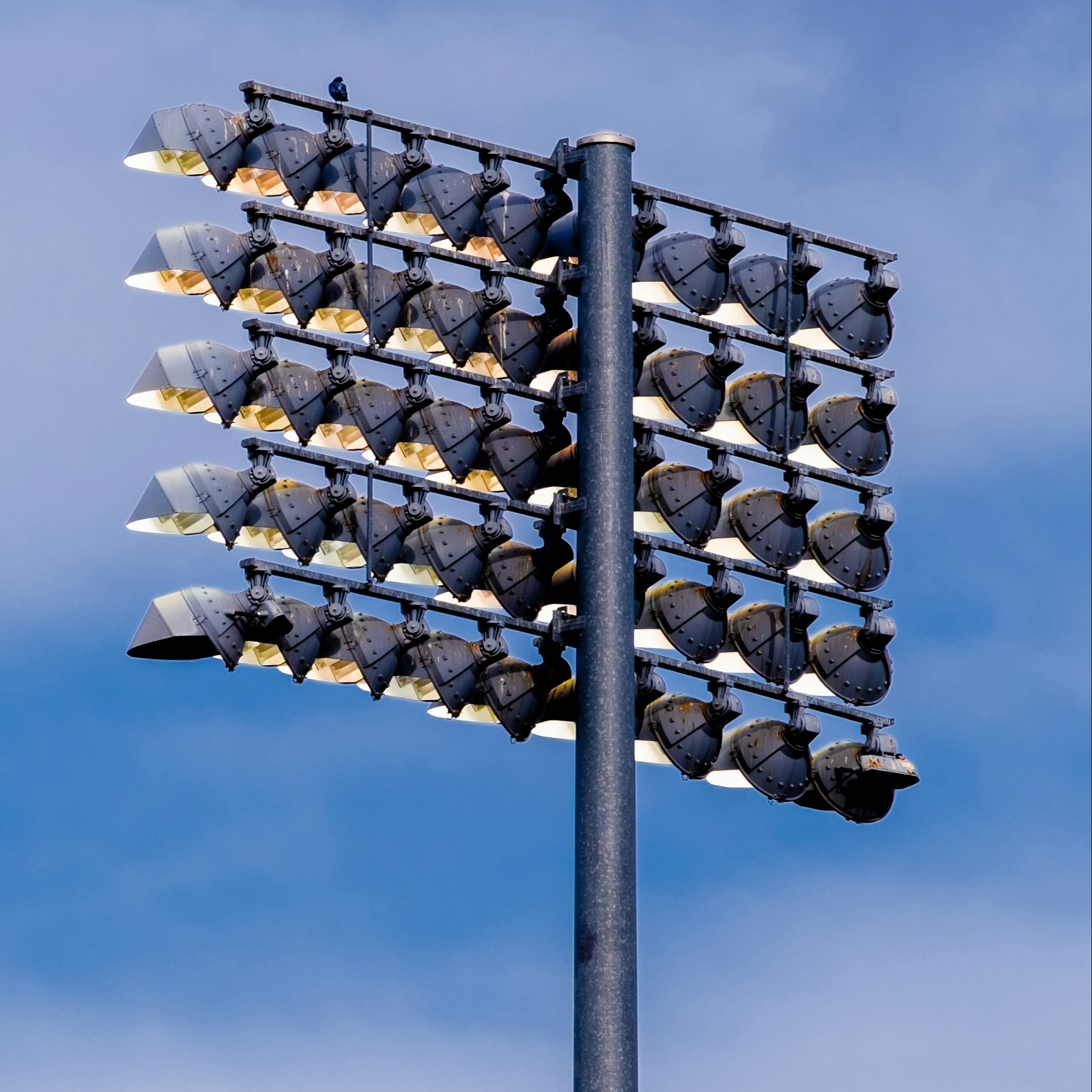 An outdoor floodlight pole with its lights on, set against a clear blue sky.