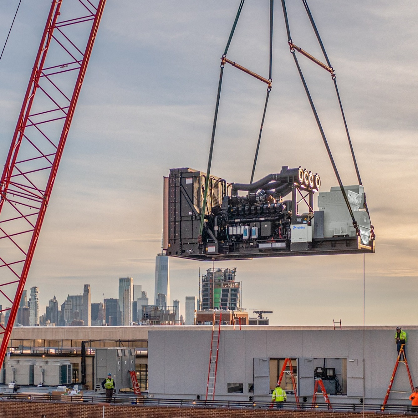 Crane lifting a diesel generator to the rooftop of a building under a warm late afternoon sky.