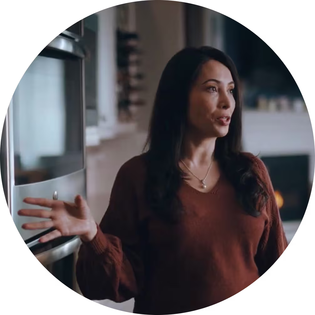 Long haired woman, wearing a brown blouse, in front of the appliances in her kitchen, talking to someone off-camera.
