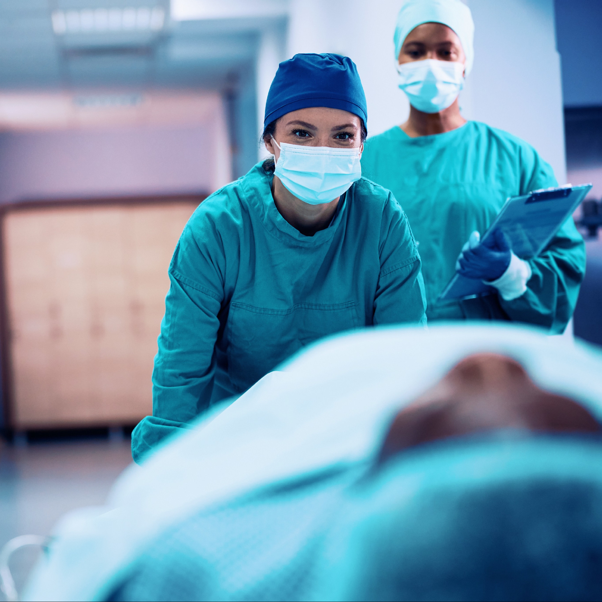 Hospital workers in scrubs and masks carefully transporting a patient on a rolling bed through a hospital hallway.