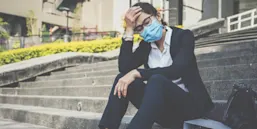 An Asian middle-aged clinician with mask sits on the steps of her hospital, a hand on her head, looking very stressed.