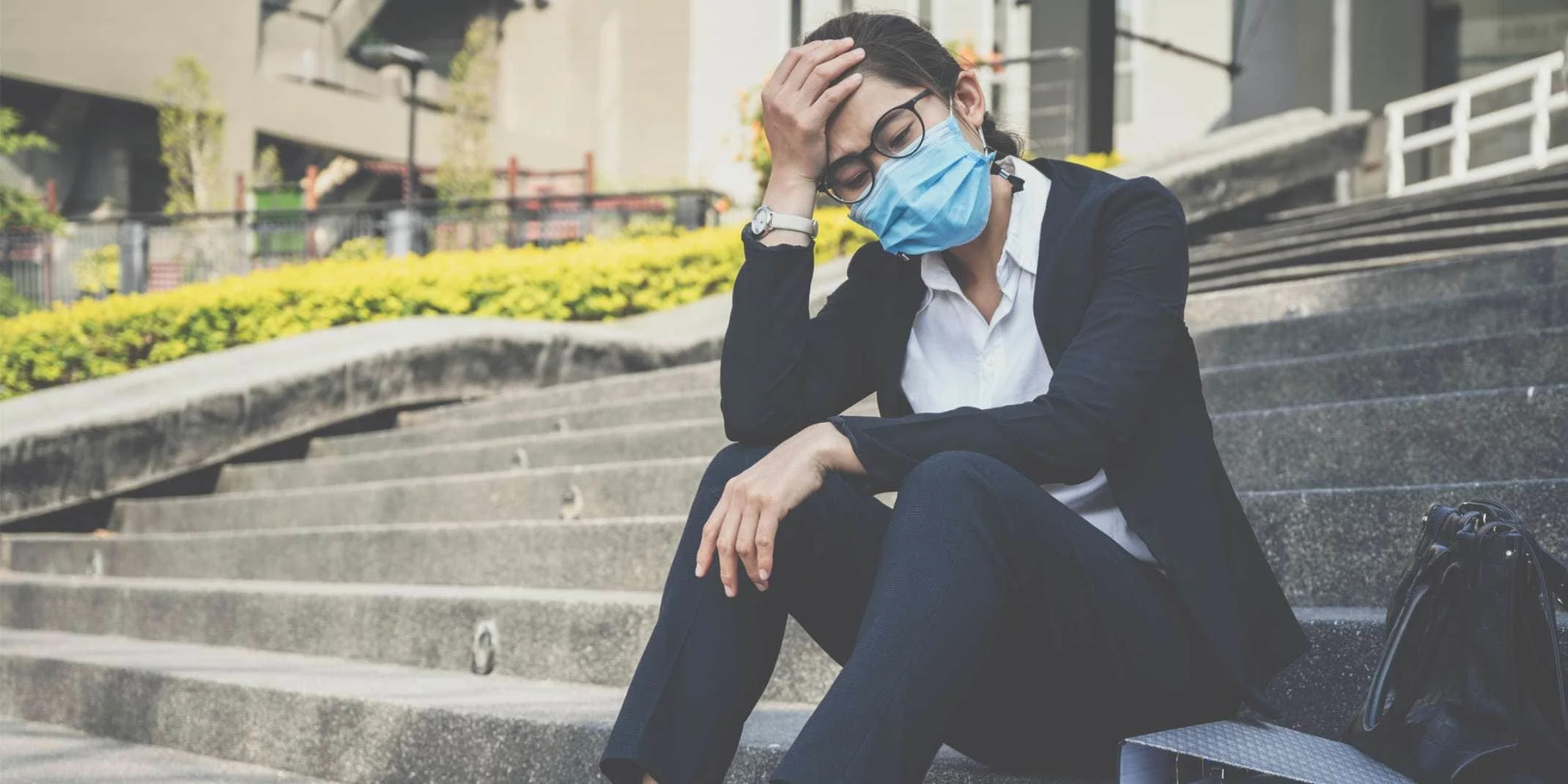 An Asian middle-aged clinician with mask sits on the steps of her hospital, a hand on her head, looking very stressed.