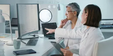 Stock photo depicting two female doctors in white coats consulting about data on a computer screen. (Source: Kobus Louw/E+ via Getty Images)
