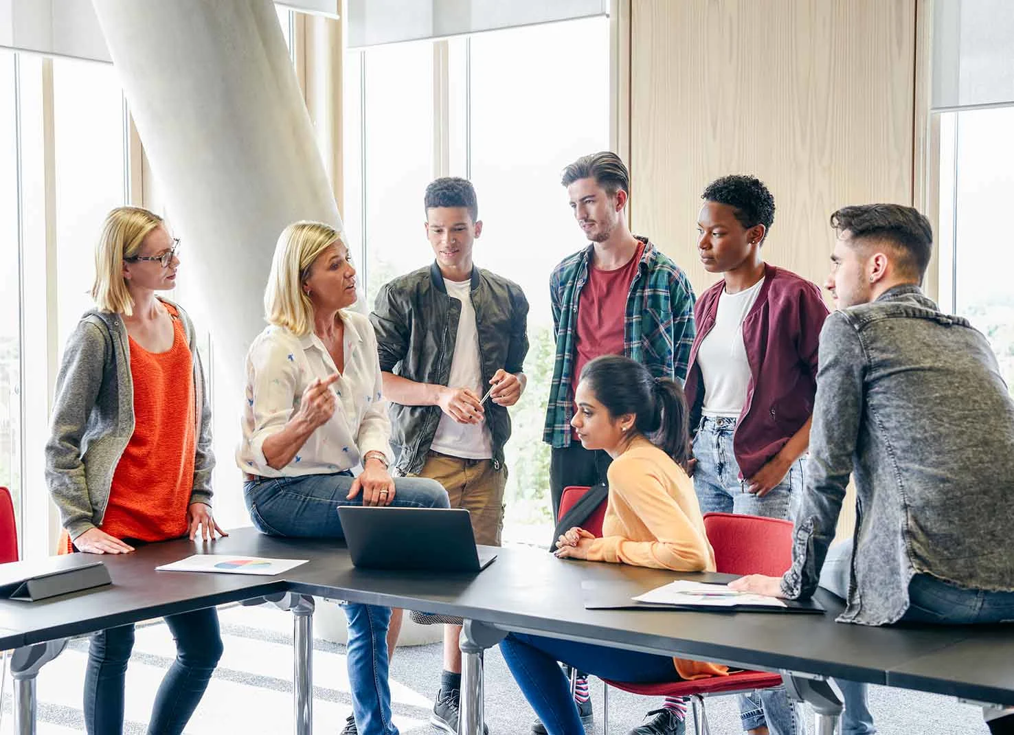 A group of students stand around a teacher explaining something on a laptop computer.