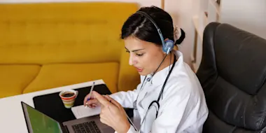 Female doctor working remotely from home using a laptop and writing notes using pen and paper