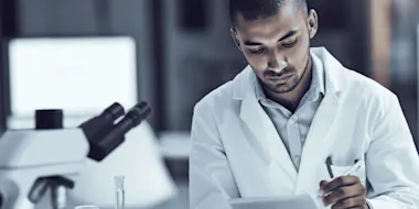 A Black male scientist, wearing a white coat and blue dress shirt, records findings on a digital tablet.