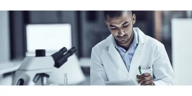 A Black male scientist, wearing a white coat and blue dress shirt, records findings on a digital tablet.