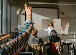 Student raising hand in class with teacher pointing at white board in front of room