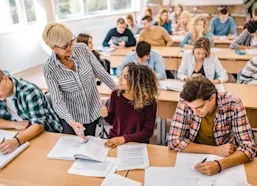 Teacher assists student with textbook in class with other students writing
