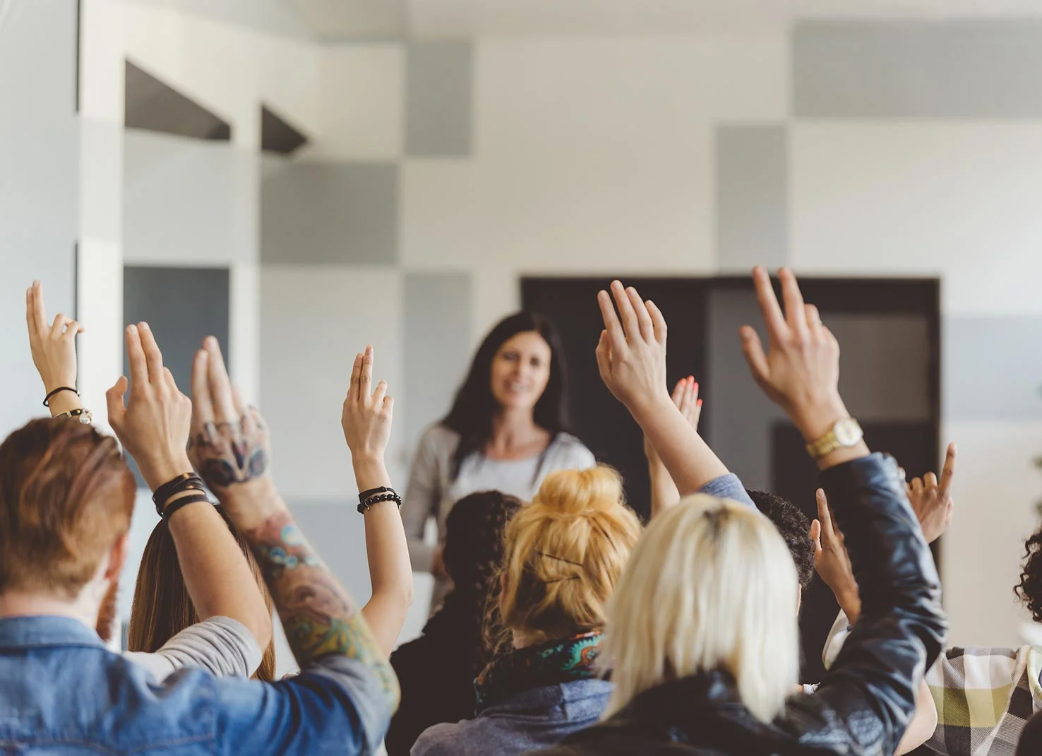 A group of students raising their hands in a classroom in front of a teacher standing.