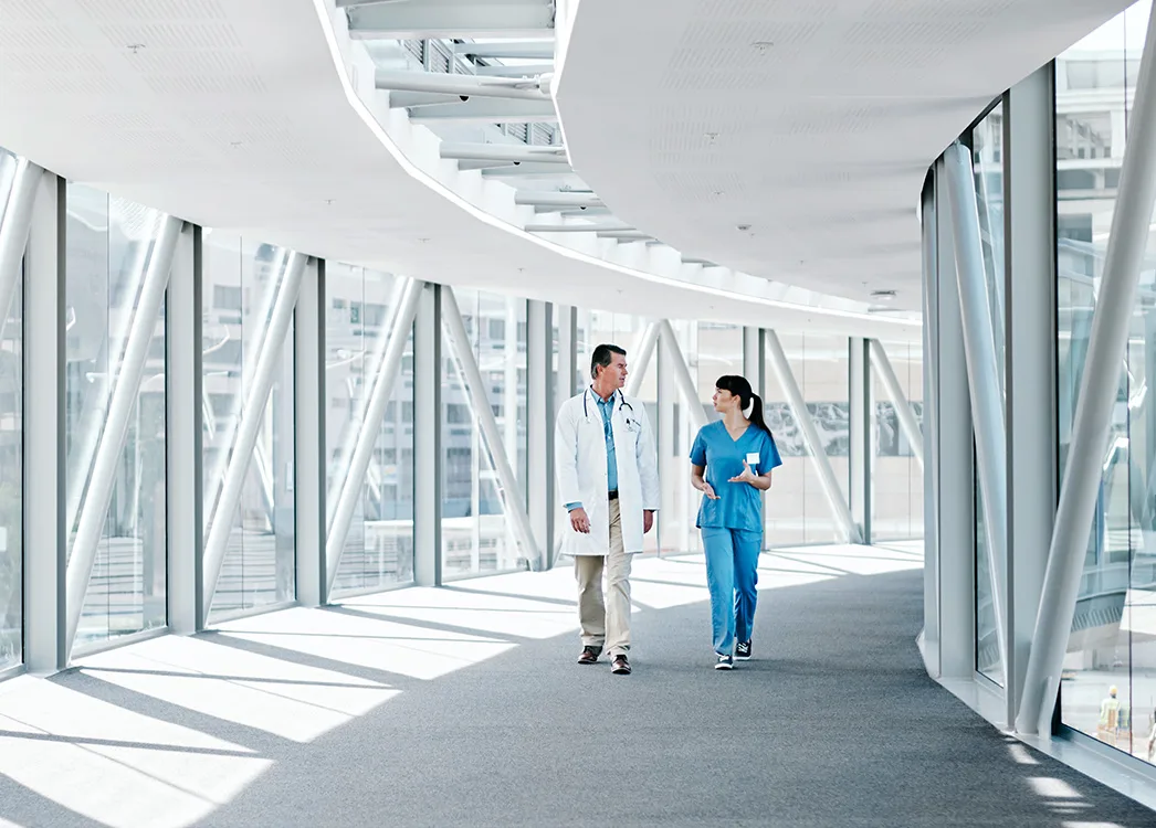 Male doctor and female nurse walk and talk down a bright hallway