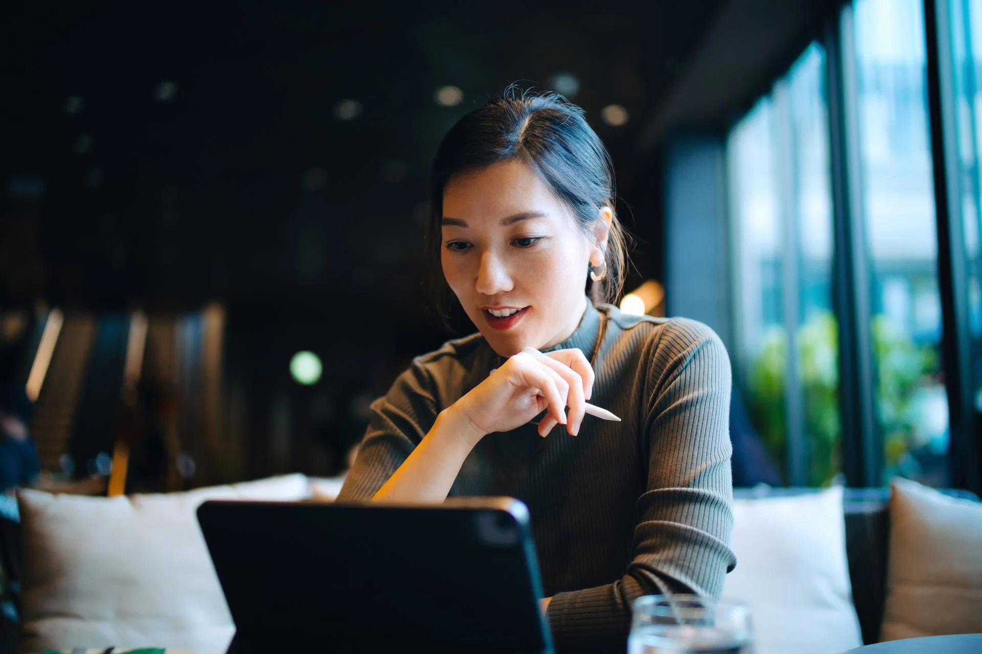 Young Asian businesswoman looking at her digital tablet while sitting at a desk in stylish co-working space and working. Successful female entrepreneur and leadership. Technology in a modern lifestyle. Working remotely with flexibility