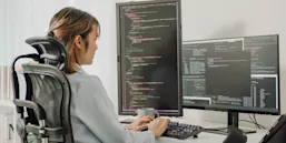 Photo of a woman at a computer with multiple screens displaying code (© istock.com/MTStock Studio)