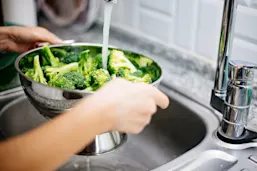 Person washing broccoli in a colander under running water
