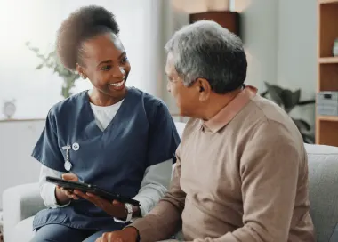 African American nurse reviews medical information on tablet with elderly male patient on couch.