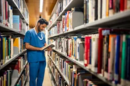 Man in blue scrubs reading a book in a library.
