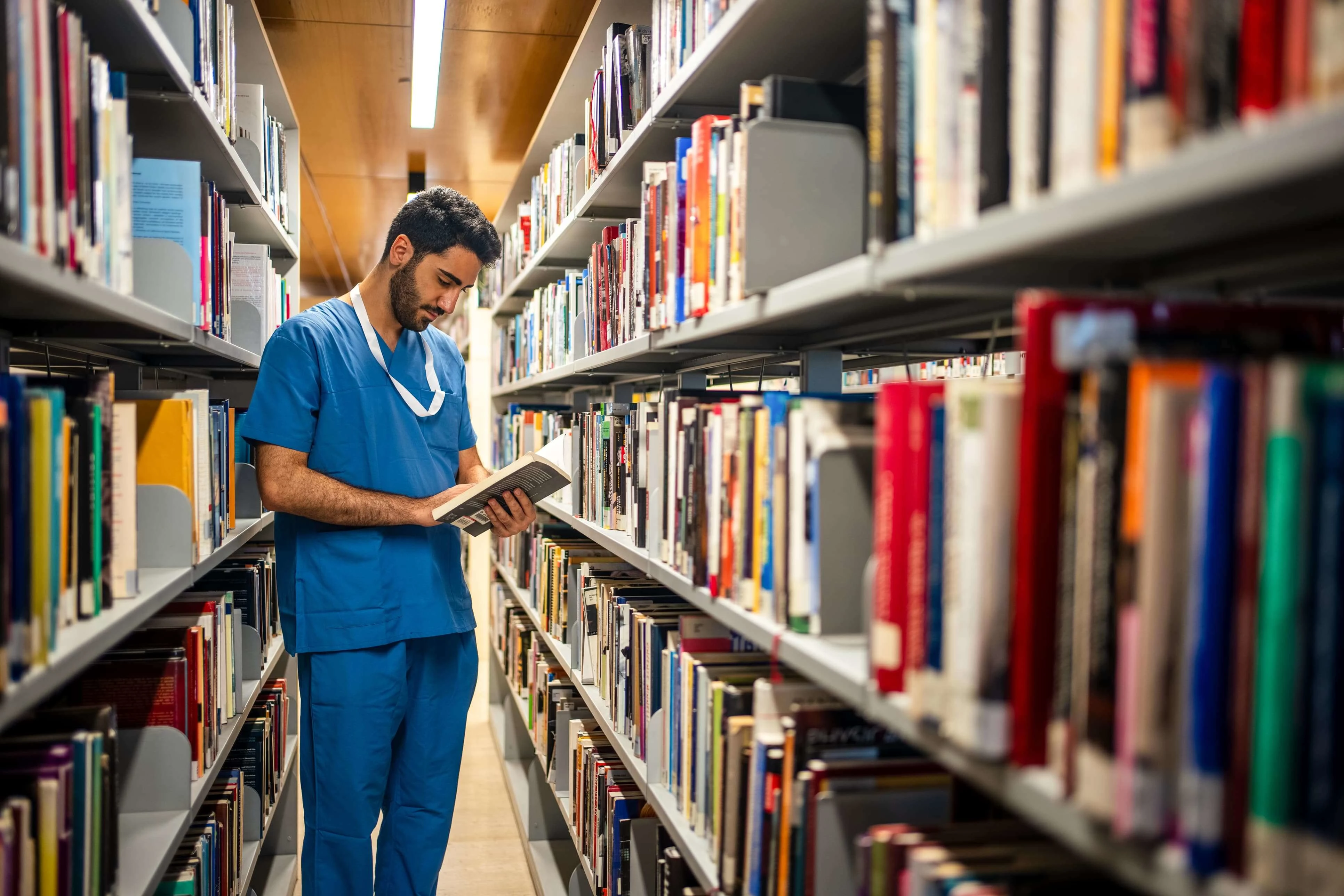 Man in blue scrubs reading a book in a library.