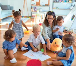 Un groupe de jeunes enfants assis par terre dans une salle de jeux qui dessinent et jouent avec des jouets, avec une femme qui s'occupe d'eux.