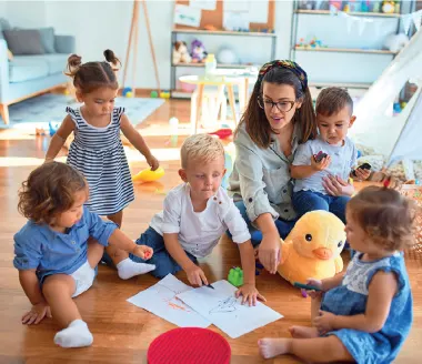 Un groupe de jeunes enfants assis par terre dans une salle de jeux qui dessinent et jouent avec des jouets, avec une femme qui s'occupe d'eux.