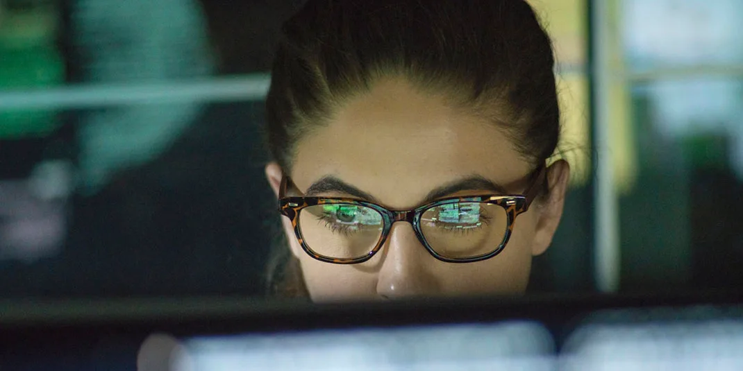 Close up stock photo of a woman surrounded by monitors and their reflections displaying scrolling text and data. (© istock.com/Laurence Dutton)