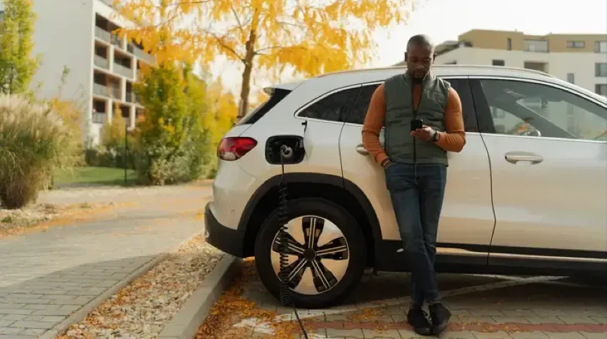A man leaning with his back against his silver Electric Vehicle. The car is plugged into an electric charger and is charging whilst the man is looking down at his mobile phone. The car is parked in a residential area with flats.