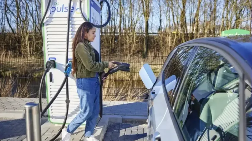 A woman is at a bp pulse charging bay and she is about to plug her Electric Vehicle in for charging. It is a bright and sunny day.