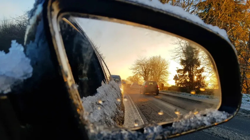 A reflection of receding traffic in a car's icy wing mirror, with frost and ice partially obscuring the view, showing blurred tail lights of vehicles that have passed and the frozen surface of the car door in the foreground.