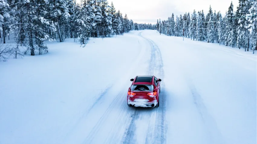 A red car driving on a trail of road, lined with trees, in the snow.