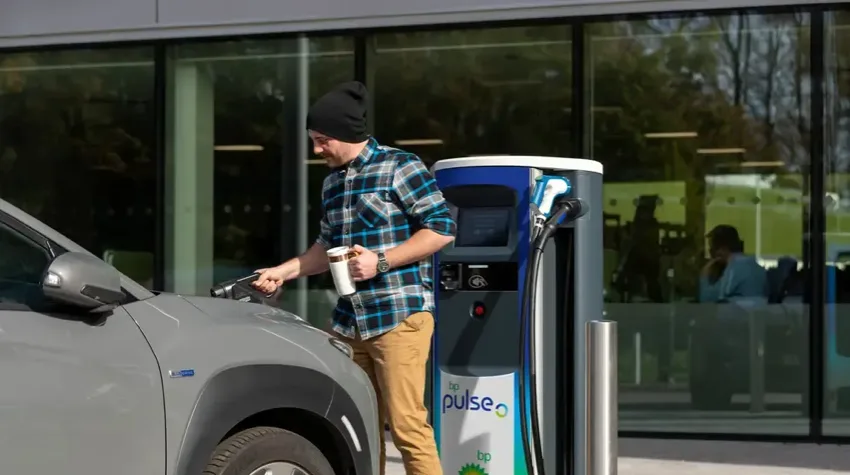 A man is standing infront of his grey EV, holding the charging lead in one hand and a coffee cup in the other. He plugging the lead from a bp pulse 50kW charger into his car. The charger is located outside modern offices with large windows.