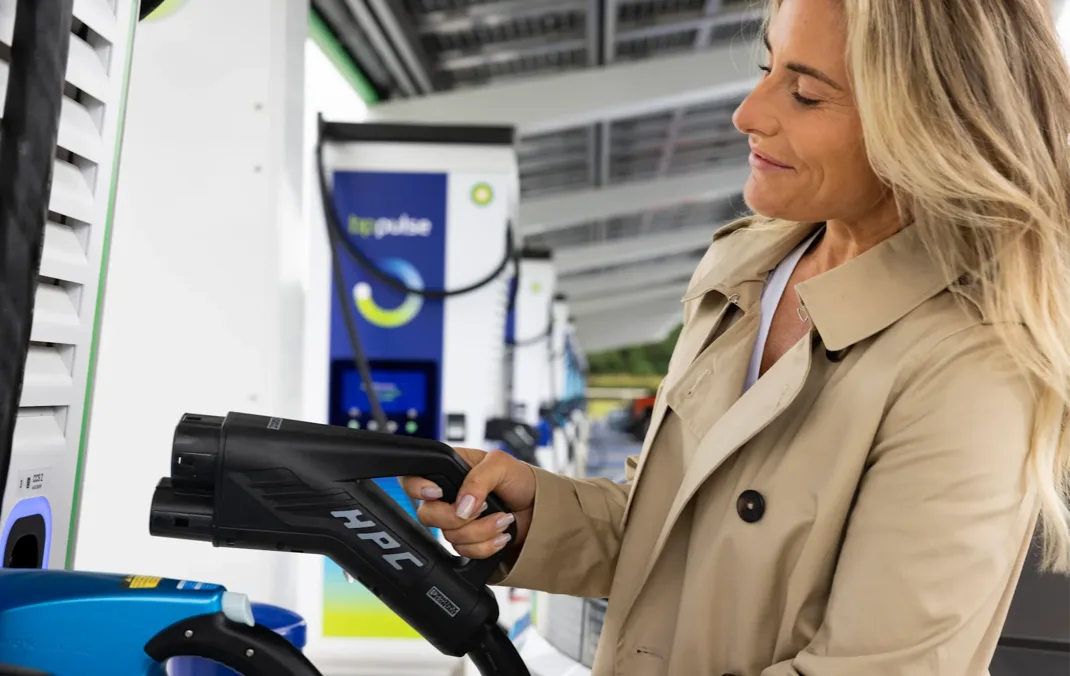 A woman wearing a light trench coat is picking up a charging plug at a bp pulse charging station.