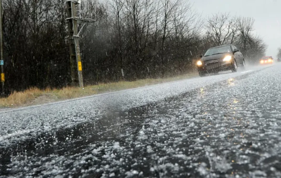 Two cars with their headlights on driving along a road during a hailstorm, with small hailstones visible bouncing off the road and a cloudy, overcast sky in the background.