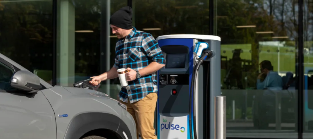 A man in a black winter hat, and flannel check shirt, is about to charge his grey EV, whilst holding a reuseable coffee mug. 