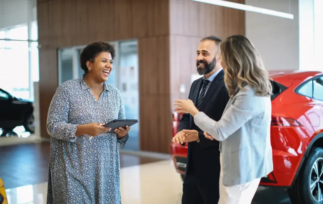 Two women and a man having a conversation in a car showroom.
