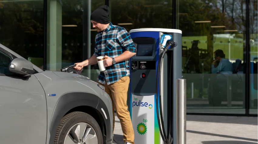 A man in a hat and a flannel shirt is charging his EV whilst holding a coffee. 