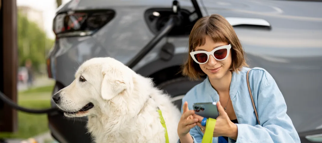A woman in sunglasses next to a light-coloured labrador, whilst her EV is charging. 
