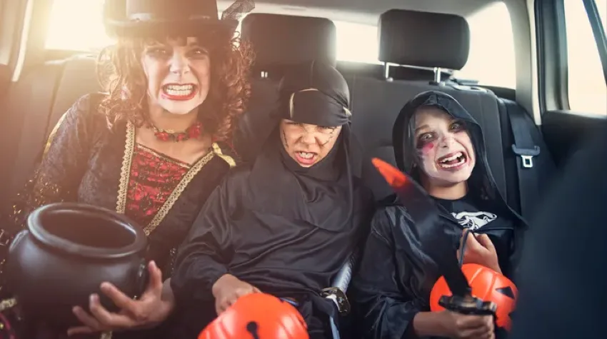 Three kids in the backseat of a car, wearing Halloween costumes and posing for the camera.