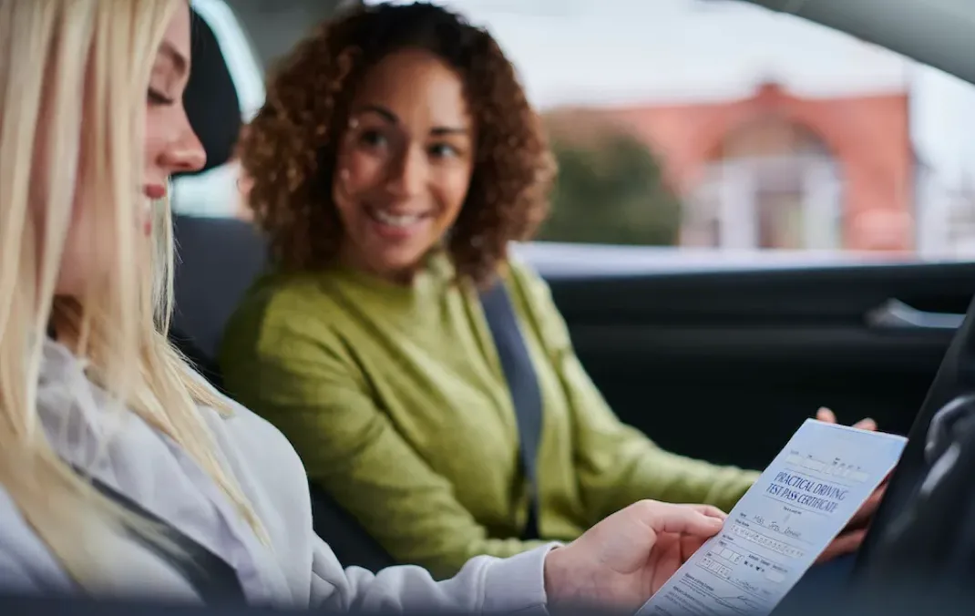A driving instructor smiles at her student who has just passed her driving test. They're sitting inside the car. 
