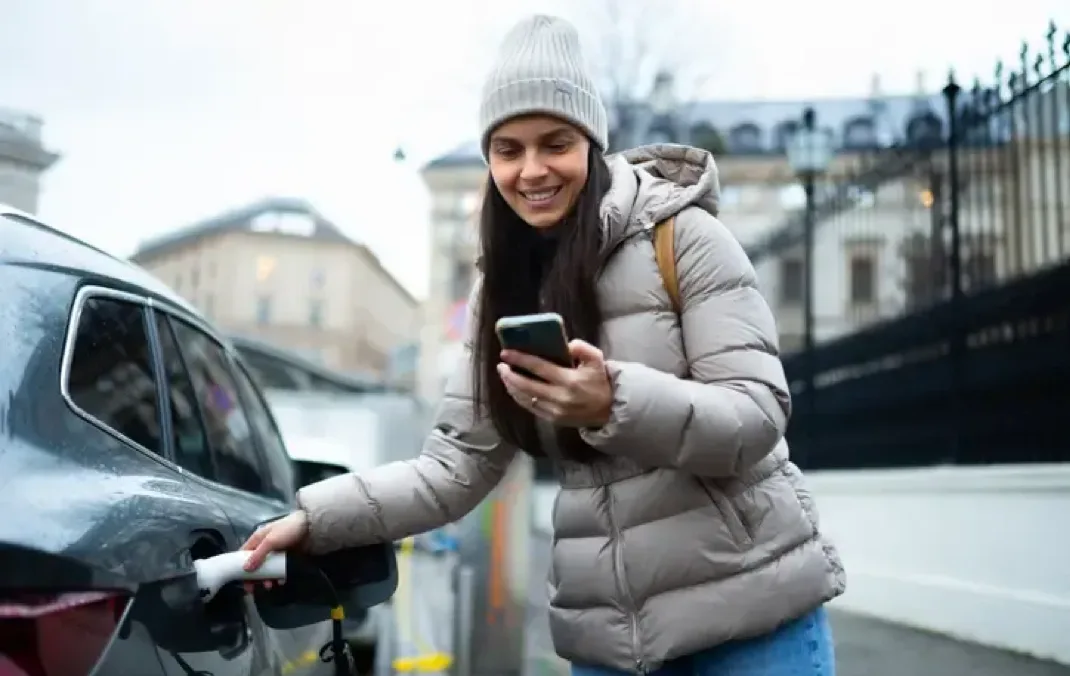 A woman wearing a padded grey coat on a dull day is charging her Electric Vehicle whilst smiling and looking down at her mobile phone. It is a dull day and black railings and tall buildings can be seen in the background.