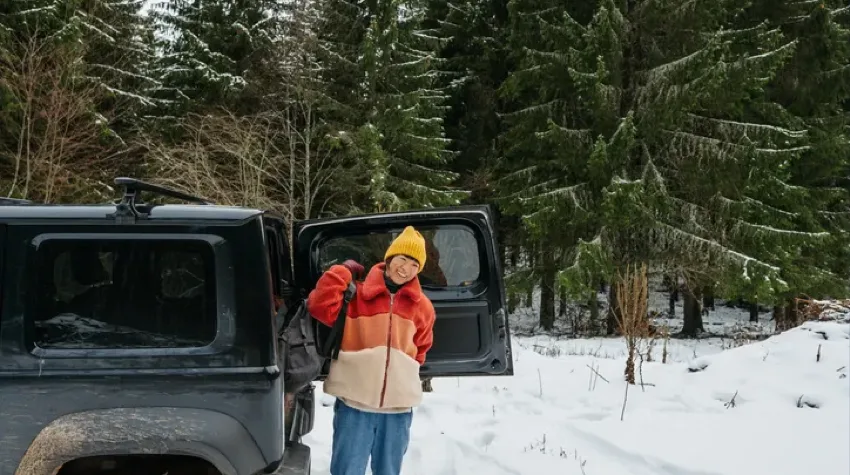 A person in a yellow winter hat and red fleece stands by the open door of their EV, in a snowy countryside location. 