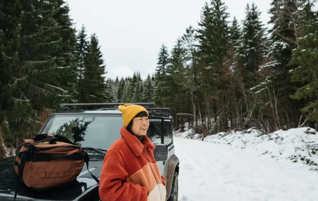 A person stands next to their EV in the snow, smiling. They are wearing a yellow winter hat and a red fleece. 