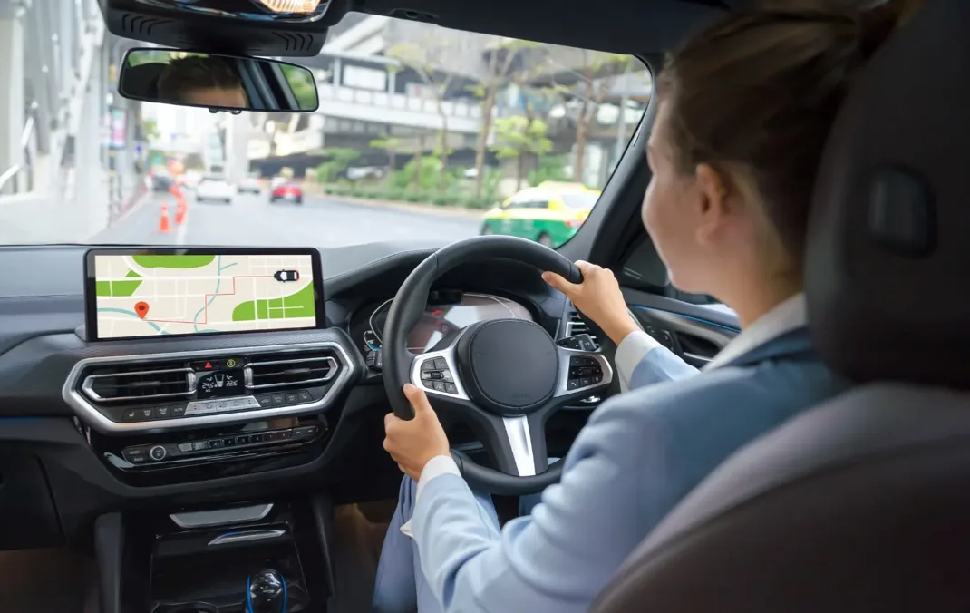 View from behind a woman's shoulder as she drives an electric vehicle with both hands on the steering wheel. The car's navigation system is visible and is displaying a route. The scene outside the windscreen shows cars driving away in the distance in a built up area.
