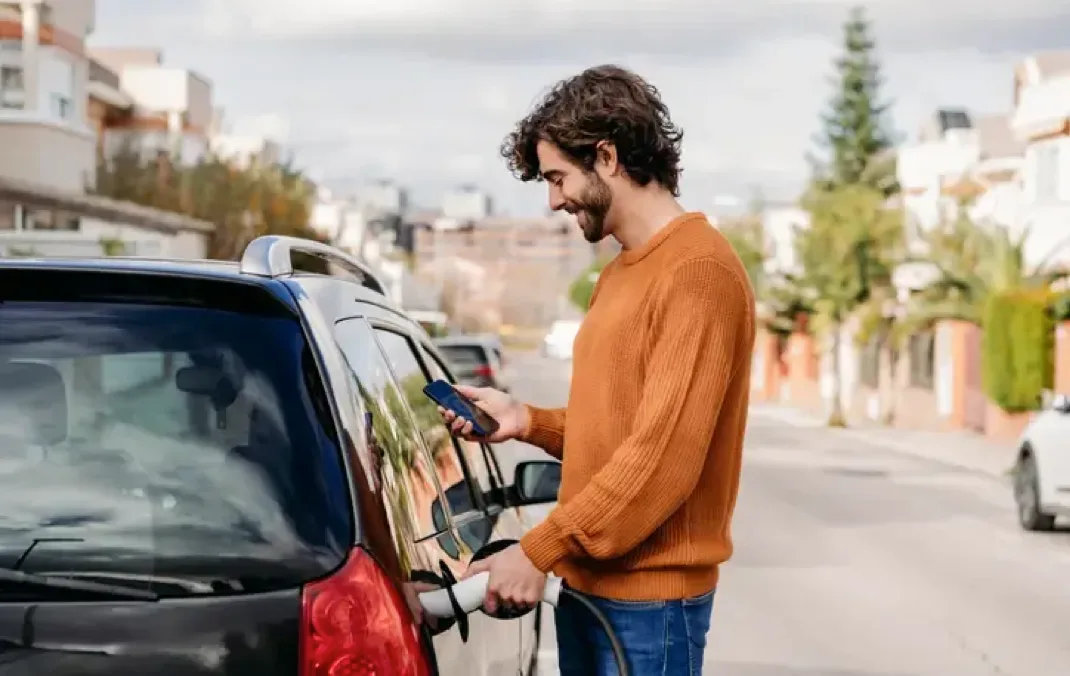 A man smiling while looking charging his electric car with one hand and holding his phone with another.