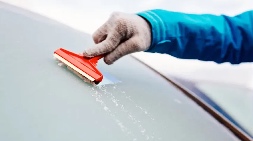A gloved hand is holding a scraper and scraping the iced winscreen of a vehicle. A blue sleeve is also visible.